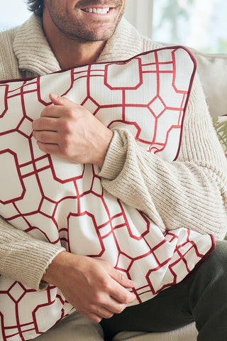 A person in a cozy beige sweater hugs the Red Holiday Lattice Oversized Pillow while sitting on a light-colored sofa, smiling gently—an ideal festive accent for your holiday decor.