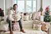 A man in a cozy sweater sits on a white sofa decorated with festive pillows, including the Bows & Mistletoe Lumbar Pillow. Small Christmas trees and wrapped gifts in the background create a cheerful holiday vibe.