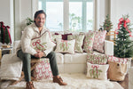 A man in a cozy sweater sits on a white sofa decorated with festive pillows, including the Bows & Mistletoe Lumbar Pillow. Small Christmas trees and wrapped gifts in the background create a cheerful holiday vibe.