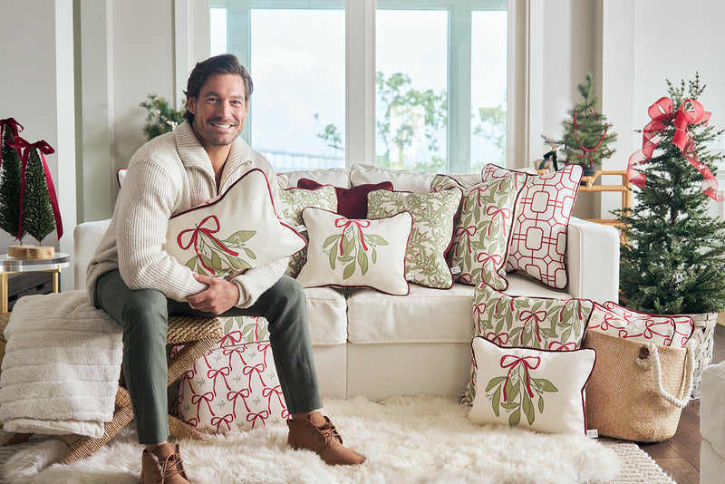 A man in a cozy sweater sits on a white sofa decorated with festive pillows, including the Bows & Mistletoe Lumbar Pillow. Small Christmas trees and wrapped gifts in the background create a cheerful holiday vibe.