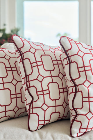 Three white throw pillows, including the Red Holiday Lattice Oversized Pillow, display a geometric red pattern and red piping on a white sofa, with soft natural light shining through a blurred window in the background.