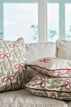 The Bows & Mistletoe Lumbar Pillow, featuring a festive mistletoe design, is displayed with two other decorative pillows on a light sofa in front of large windows overlooking a blurred outdoor view.