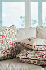 The Bows & Mistletoe Lumbar Pillow, featuring a festive mistletoe design, is displayed with two other decorative pillows on a light sofa in front of large windows overlooking a blurred outdoor view.