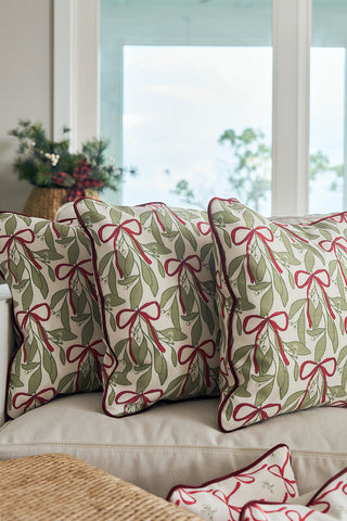 The Oh Mistletoe Oversized Pillow, featuring a green and red mistletoe pattern, sits on a light sofa by a bright window, adding a cozy and festive touch to the seasonal decor.