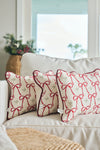 Two white Bows & Mistletoe Lumbar Pillows featuring red ribbon bow designs sit on a light sofa, with a woven basket of greenery and large windows providing natural light in the background.