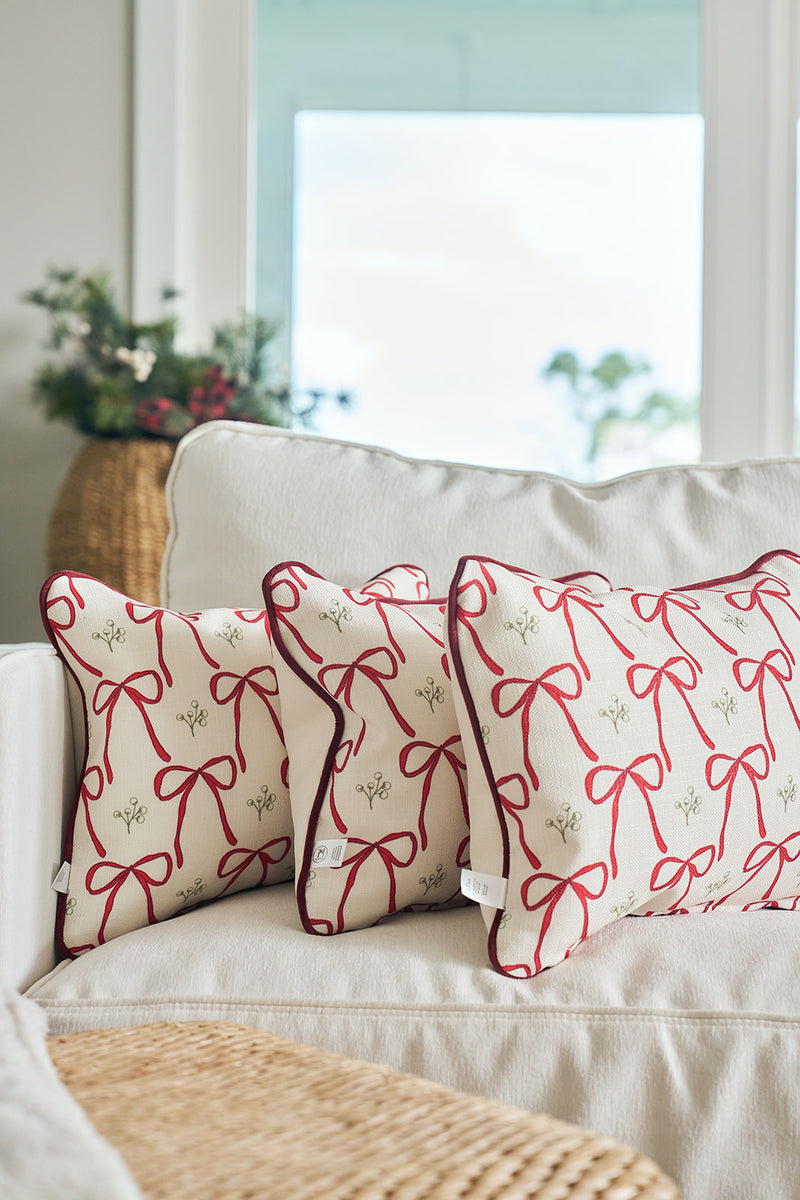 Two white Bows & Mistletoe Lumbar Pillows featuring red ribbon bow designs sit on a light sofa, with a woven basket of greenery and large windows providing natural light in the background.