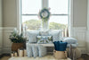 A cozy bench by a window features the Silver Bells Pillow, blue and white patterned pillows, twill woven pillow, gift boxes, a wreath, small presents, a potted plant, and blue-gold baskets on a fluffy rug.