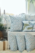 A cozy seating area features the Silver Bells Pillow along with white and blue twill woven pillows, all adorned with snowflakes and holiday motifs, set among festive greenery and soft, fluffy textures.