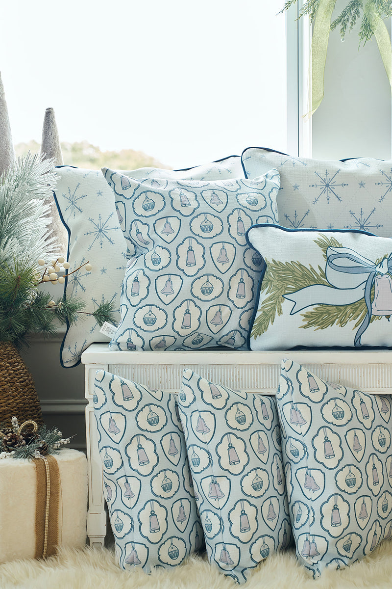 A cozy seating area features the Silver Bells Pillow along with white and blue twill woven pillows, all adorned with snowflakes and holiday motifs, set among festive greenery and soft, fluffy textures.