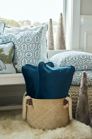 A cozy interior features the Holiday Swag Extended Lumbar Pillow with blue and white patterns on a bench, two solid blue pillows in a woven basket, and soft gray cone-shaped decor on a fluffy rug by the window.