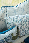 The Silver Bells Pillow, featuring blue and white designs of snowflakes, bells, and a festive bow with greenery on twill fabric, sits with two other decorative holiday pillows on a white surface against a softly blurred background.