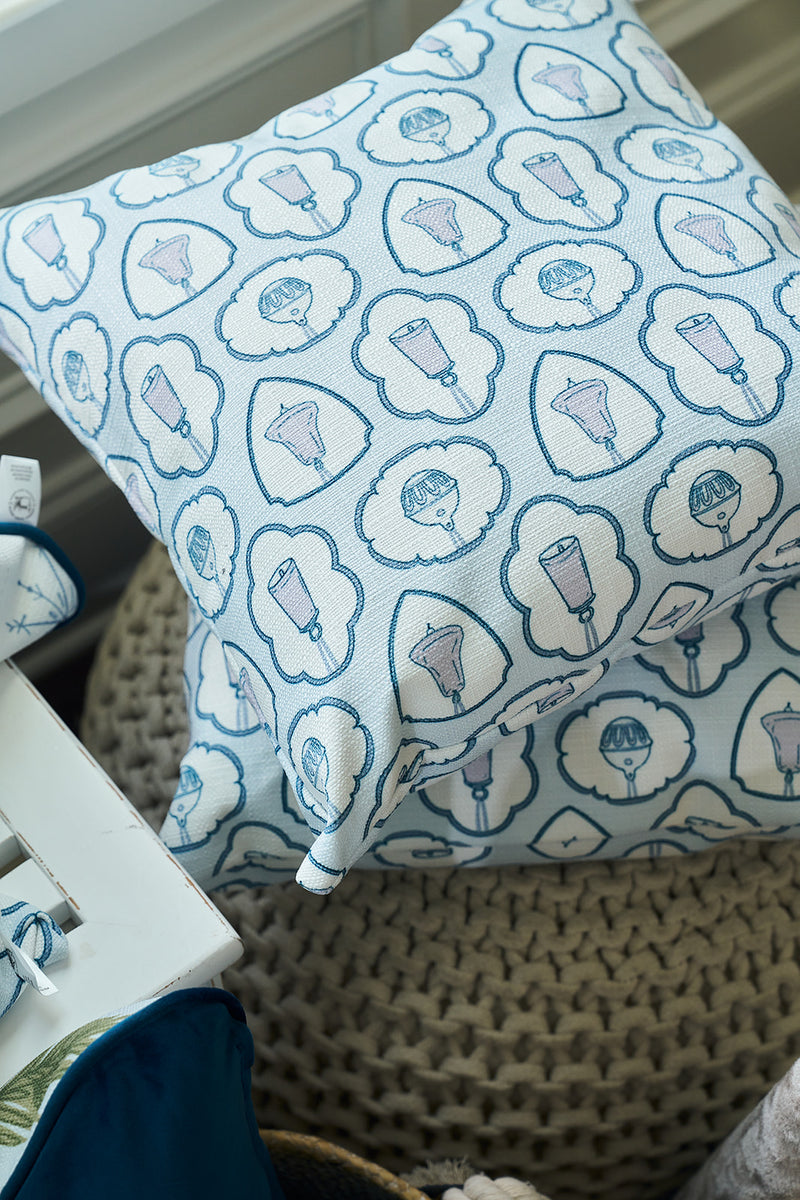 The Silver Bells Pillow, with its blue and white twill weave and holiday-inspired patterns, rests on a knitted pouf near a window, making it the perfect festive accent for any cozy space.