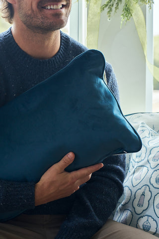 A person in a blue sweater smiles on a sofa, holding the Holiday Swag Extended Lumbar Pillow. Patterned cushions and a window with greenery appear in the background.