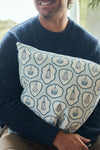 A smiling person in a blue sweater sits indoors by a window, holding the Silver Bells Pillow, which features twill woven designs of bells, shields, and circular shapes in light blue and white tones.