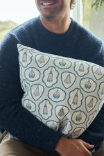 A smiling person in a blue sweater sits indoors by a window, holding the Silver Bells Pillow, which features twill woven designs of bells, shields, and circular shapes in light blue and white tones.
