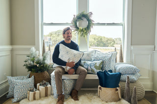 A man sits on a window bench hugging the Luxe Snowflakes Oversized Pillow, surrounded by winter-themed pillows, wrapped gifts, baskets, and a small decorated pine tree. A green wreath is displayed in the window behind him.