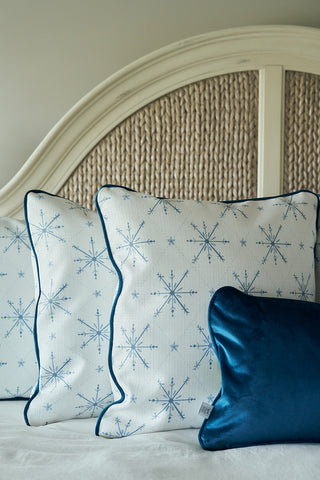 Three decorative pillows rest on a bed with a woven, arched headboard: two Luxe Snowflakes Oversized Pillows with blue snowflake designs, and one smaller shiny blue Christmas pillow from Sewing Down South.