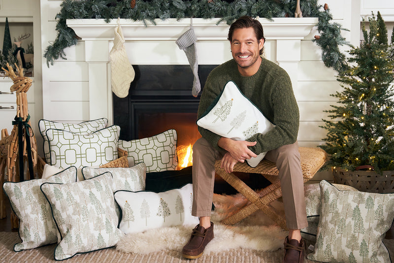 A man in a green sweater sits by a decorated fireplace with white Christmas pillows and the Row of Trees Extended Lumbar Pillow. Stockings hang overhead, while holiday greenery and a small lit tree add festive charm nearby.