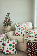 A cozy armchair features a Classic Ornaments Pillow with colorful ornament designs, paired with another holiday pillow. Extra coordinating pillows are stacked on a pouf next to a small Christmas tree in a basket.