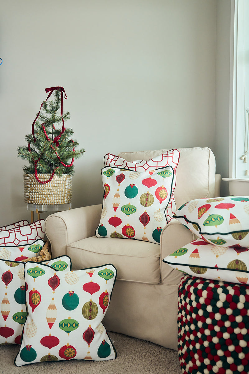 A cozy armchair features a Classic Ornaments Pillow with colorful ornament designs, paired with another holiday pillow. Extra coordinating pillows are stacked on a pouf next to a small Christmas tree in a basket.