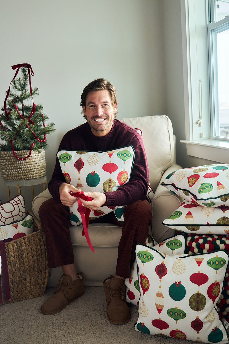 A smiling man in a maroon sweater sits in an armchair hugging a Classic Ornaments Pillow, surrounded by festive throw pillows and a small decorated Christmas tree.