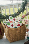 A hand places the Classic Ornaments Pillow, featuring colorful ornament designs, into a woven basket on a garland-adorned porch. Other decorative pillows fill the basket, with a railing and greenery in the background.