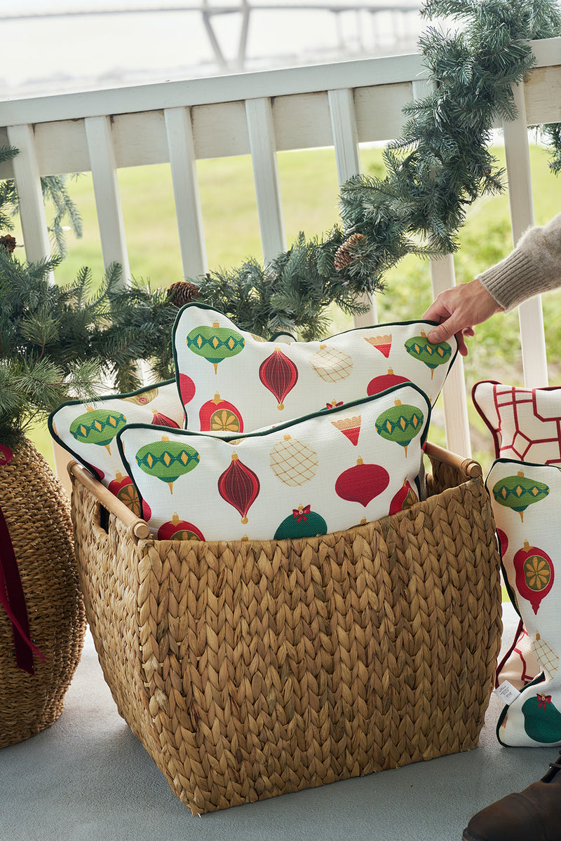 A hand places the Classic Ornaments Pillow, featuring colorful ornament designs, into a woven basket on a garland-adorned porch. Other decorative pillows fill the basket, with a railing and greenery in the background.