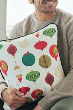 A smiling man in a beige sweater holds the Classic Ornaments Pillow, which features colorful holiday ornament patterns with classic Christmas designs in red, green, and gold.