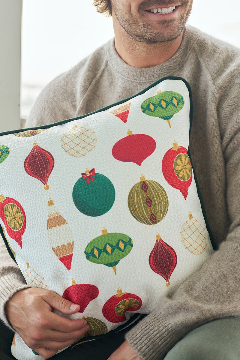 A smiling man in a beige sweater holds the Classic Ornaments Pillow, which features colorful holiday ornament patterns with classic Christmas designs in red, green, and gold.