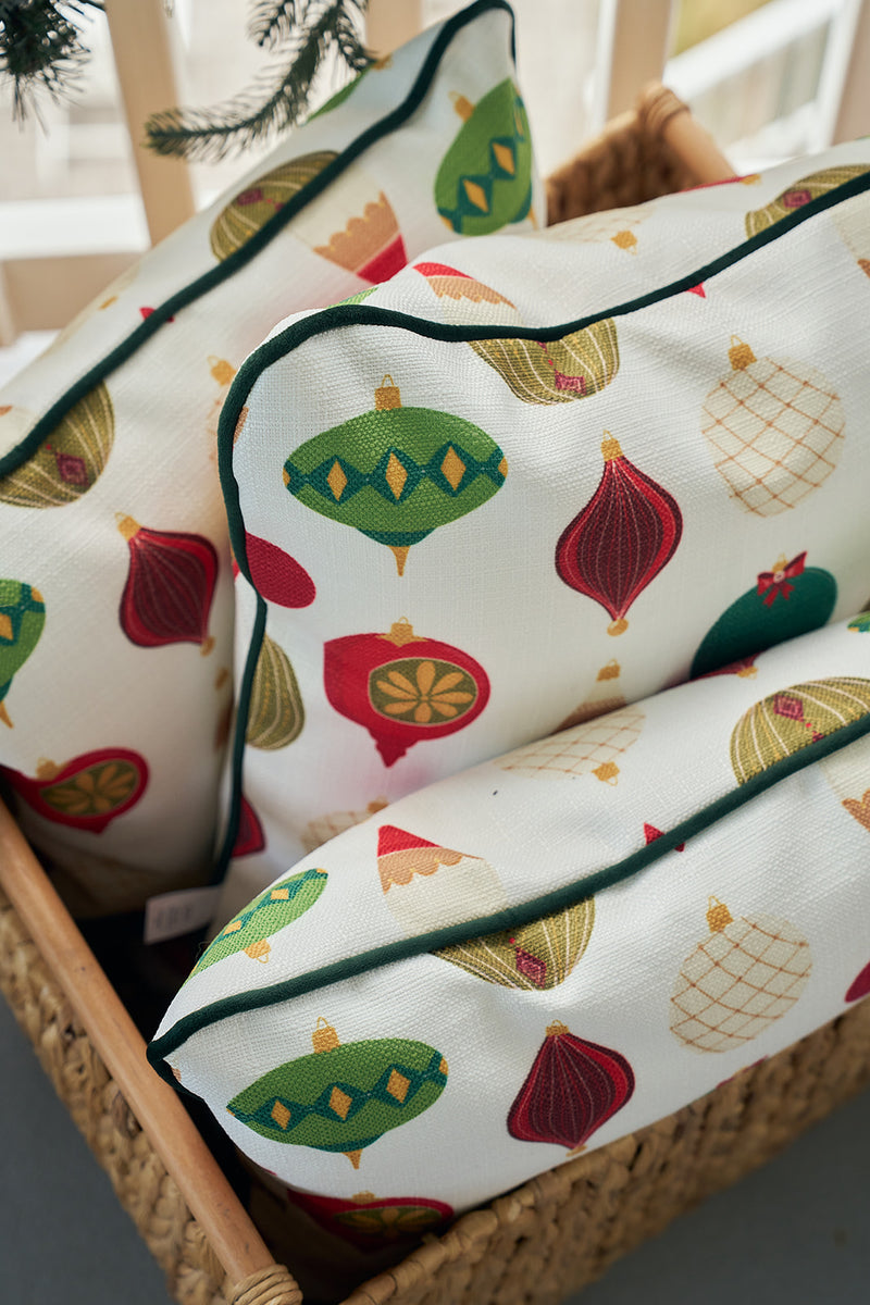Three white Classic Ornaments Pillows, featuring colorful Christmas ornament patterns and green piping, are arranged in a woven basket. Pine branches above complete the festive holiday scene.