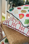 A Classic Ornaments Pillow with festive designs tops a red geometric pillow on a wicker chair, with pine branches and ornaments enhancing the holiday display.