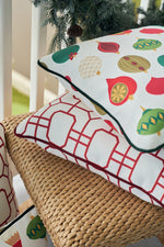 A Classic Ornaments Pillow with festive designs tops a red geometric pillow on a wicker chair, with pine branches and ornaments enhancing the holiday display.