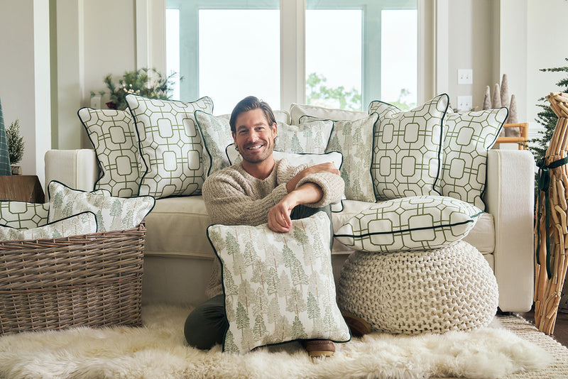 A man sits on the floor in front of a white couch with festive pillows, smiling as he holds the Christmas Tree Farm Pillow. The bright living room is filled with cozy holiday decor and patterned cushions.