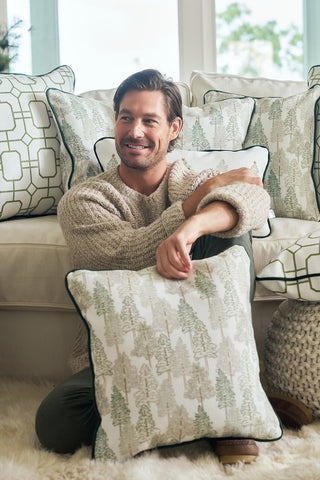 A man with brown hair, dressed in a beige sweater and green pants, sits on a fluffy rug holding the Christmas Tree Farm Pillow. Behind him, a white couch is decorated with several festive holiday pillows.