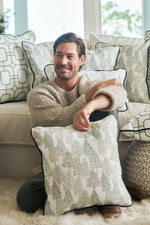 A man with brown hair, dressed in a beige sweater and green pants, sits on a fluffy rug holding the Christmas Tree Farm Pillow. Behind him, a white couch is decorated with several festive holiday pillows.
