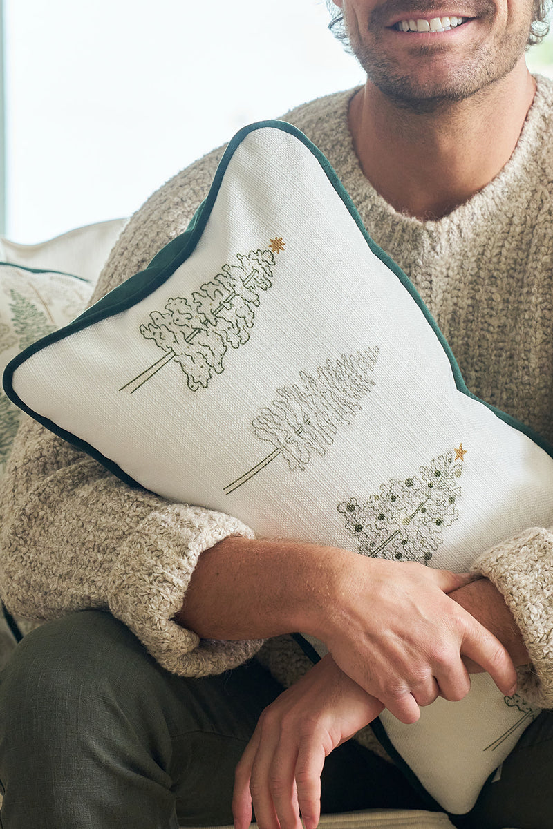 A person in a cozy beige sweater holds the Row of Trees Extended Lumbar Pillow—a white holiday pillow with embroidered green trees, gold stars, and dark green piping around the edges.