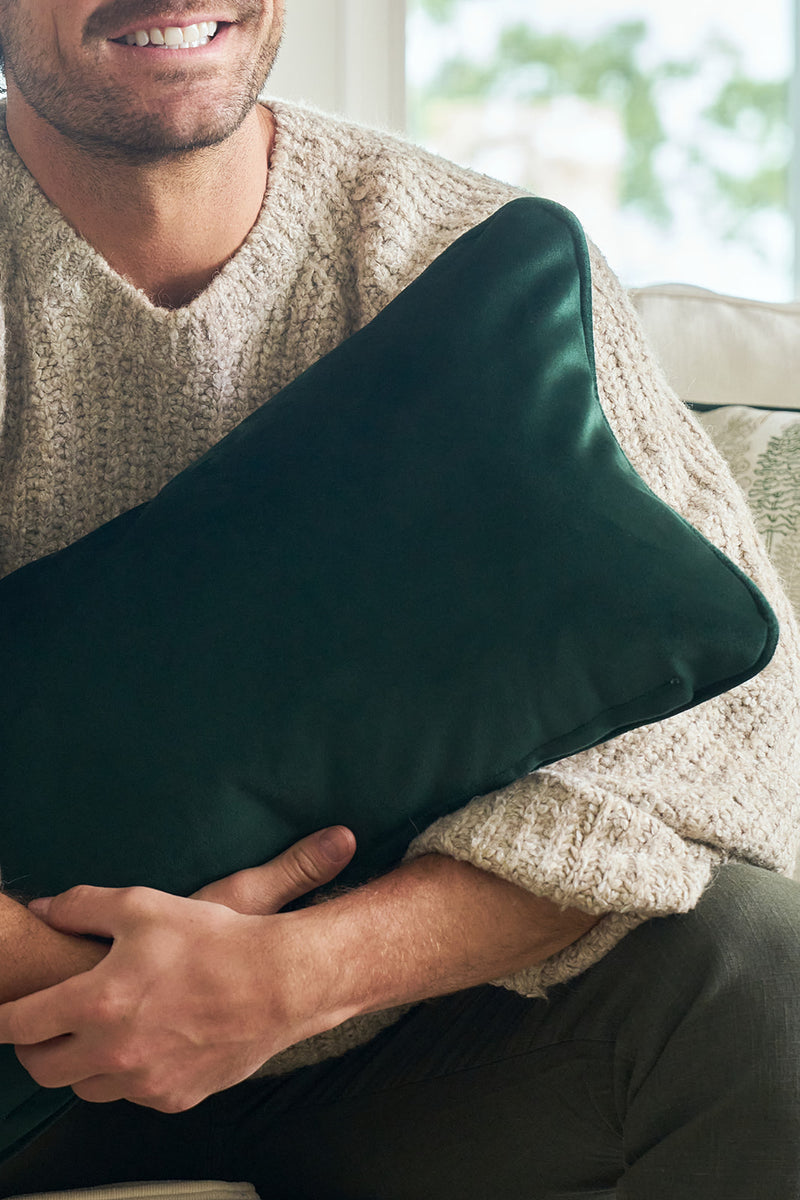 A person in a beige knit sweater sits on a couch, smiling and holding the Row of Trees Extended Lumbar Pillow against their chest. Only their lower face and torso are visible.