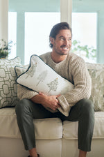 A man with brown hair and a beard, in a beige sweater and gray pants, smiles on a white couch while hugging a Christmas tree pillow. Festive geometric pillows and the Row of Trees Extended Lumbar Pillow are behind him.
