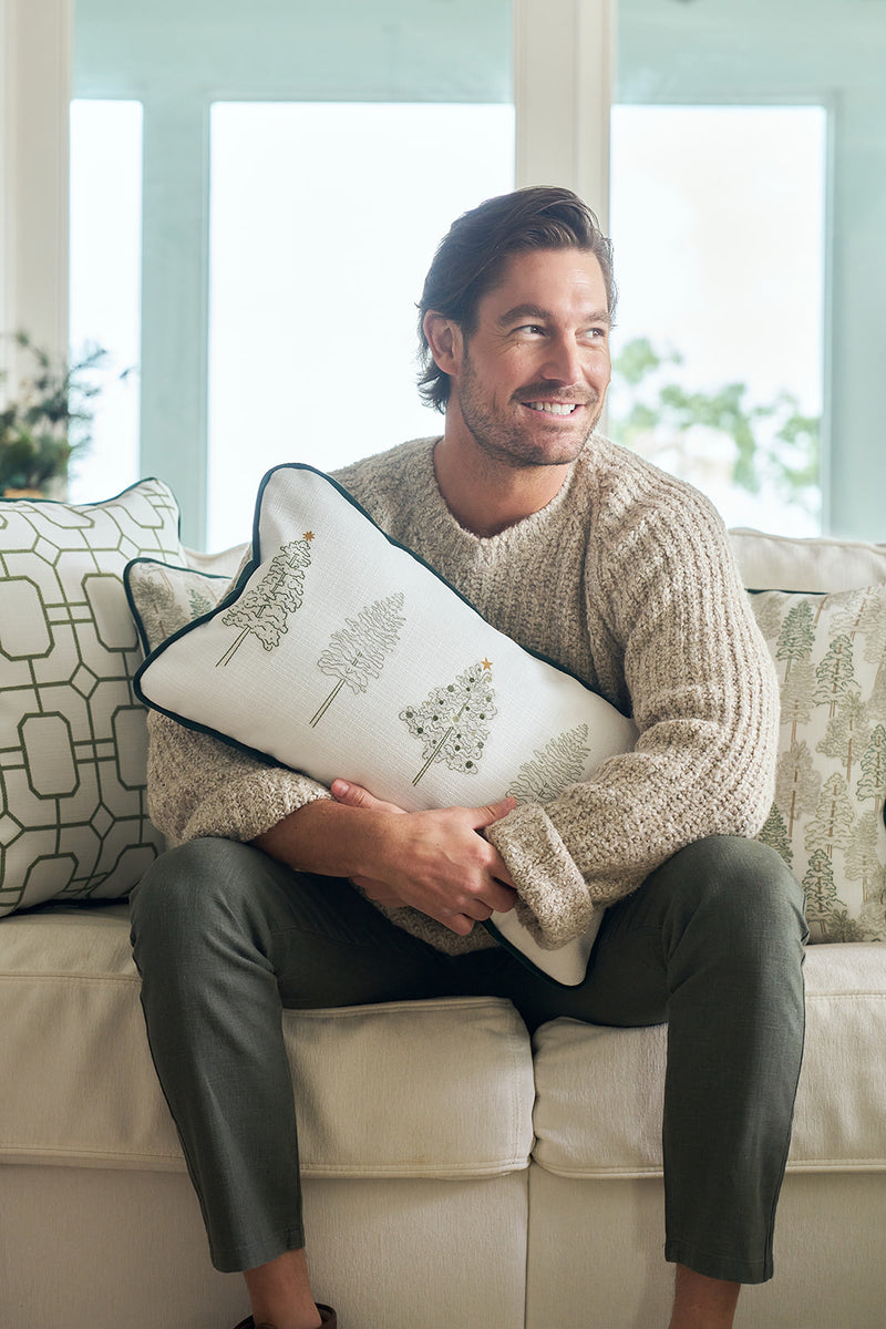 A man with brown hair and a beard, in a beige sweater and gray pants, smiles on a white couch while hugging a Christmas tree pillow. Festive geometric pillows and the Row of Trees Extended Lumbar Pillow are behind him.