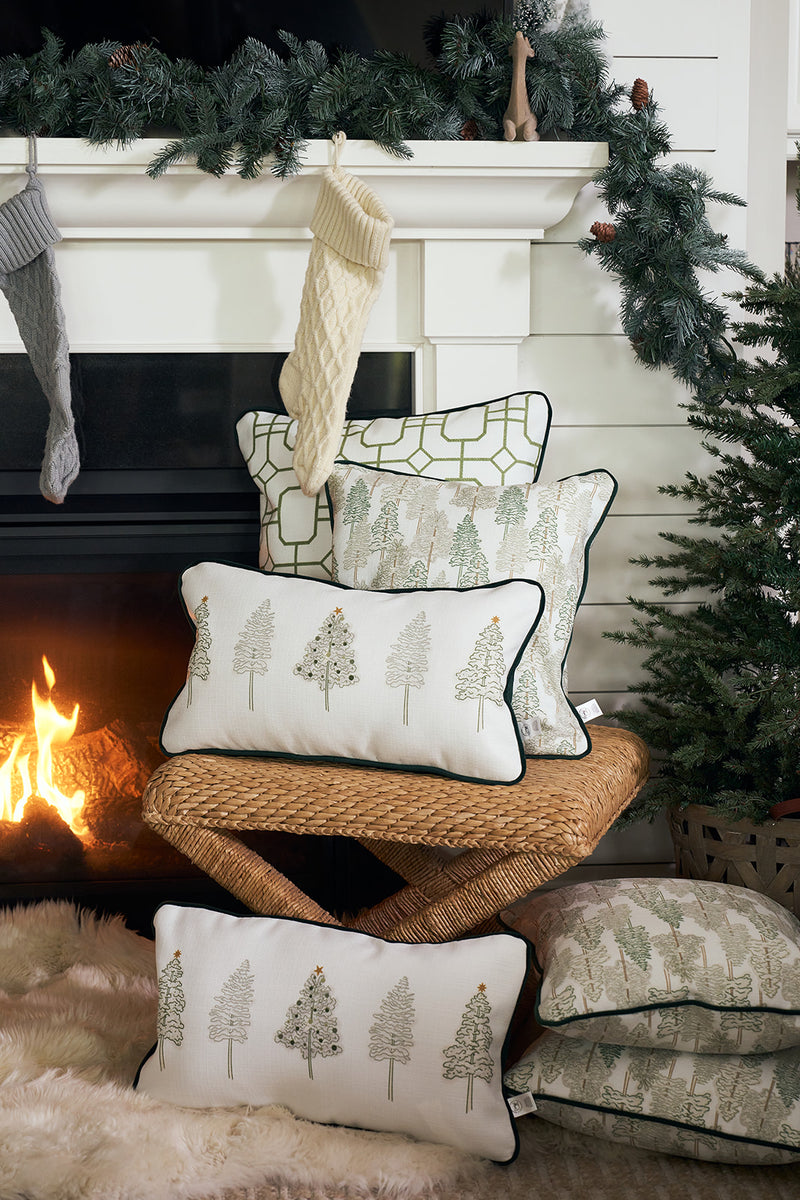 A cozy fireplace scene features a wicker bench stacked with white pillows, including the Row of Trees Extended Lumbar Pillow, plus a small Christmas tree, stockings on the mantle, pine garland, and a fur rug.