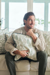 A bearded man with dark hair smiles on a couch, hugging the Christmas Tree Farm Pillow. He wears a beige knit sweater and green pants, surrounded by other patterned pillows for a cozy holiday feel.