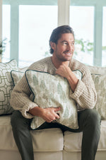 A bearded man with dark hair smiles on a couch, hugging the Christmas Tree Farm Pillow. He wears a beige knit sweater and green pants, surrounded by other patterned pillows for a cozy holiday feel.
