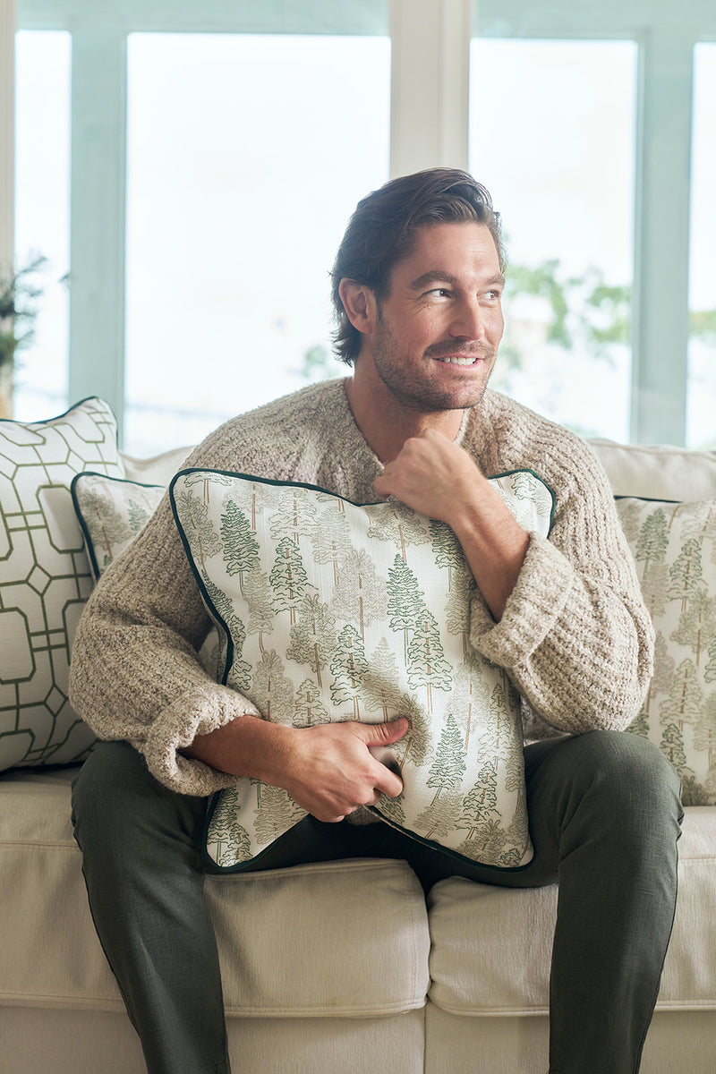 A bearded man with dark hair smiles on a couch, hugging the Christmas Tree Farm Pillow. He wears a beige knit sweater and green pants, surrounded by other patterned pillows for a cozy holiday feel.