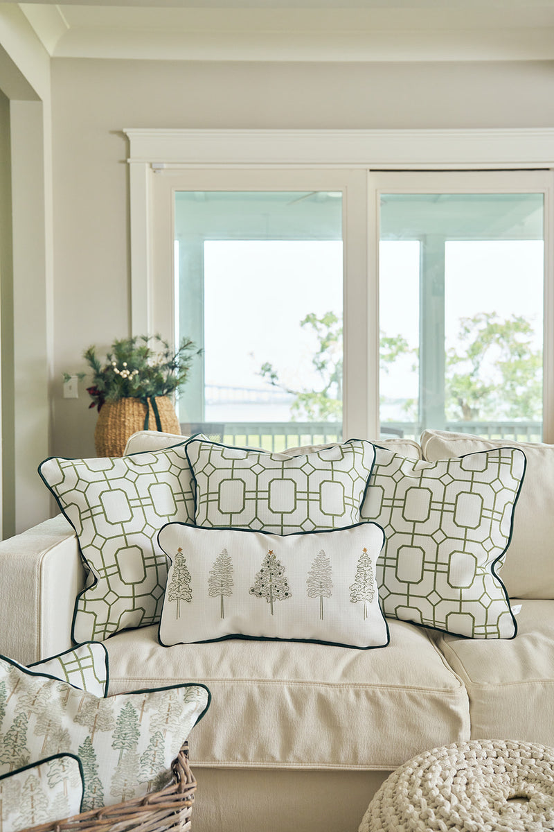 A bright, cozy living room with a white sofa adorned with three green geometric pillows and the Row of Trees Extended Lumbar Pillow, set near a large window showcasing a scenic outdoor view.