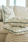 A Christmas Tree Farm Pillow rests on a knitted ottoman in front of a beige sofa with matching pillows, creating a cozy and inviting living room atmosphere.