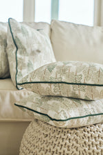 A Christmas Tree Farm Pillow rests on a knitted ottoman in front of a beige sofa with matching pillows, creating a cozy and inviting living room atmosphere.