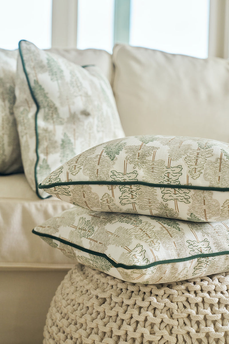 A Christmas Tree Farm Pillow rests on a knitted ottoman in front of a beige sofa with matching pillows, creating a cozy and inviting living room atmosphere.