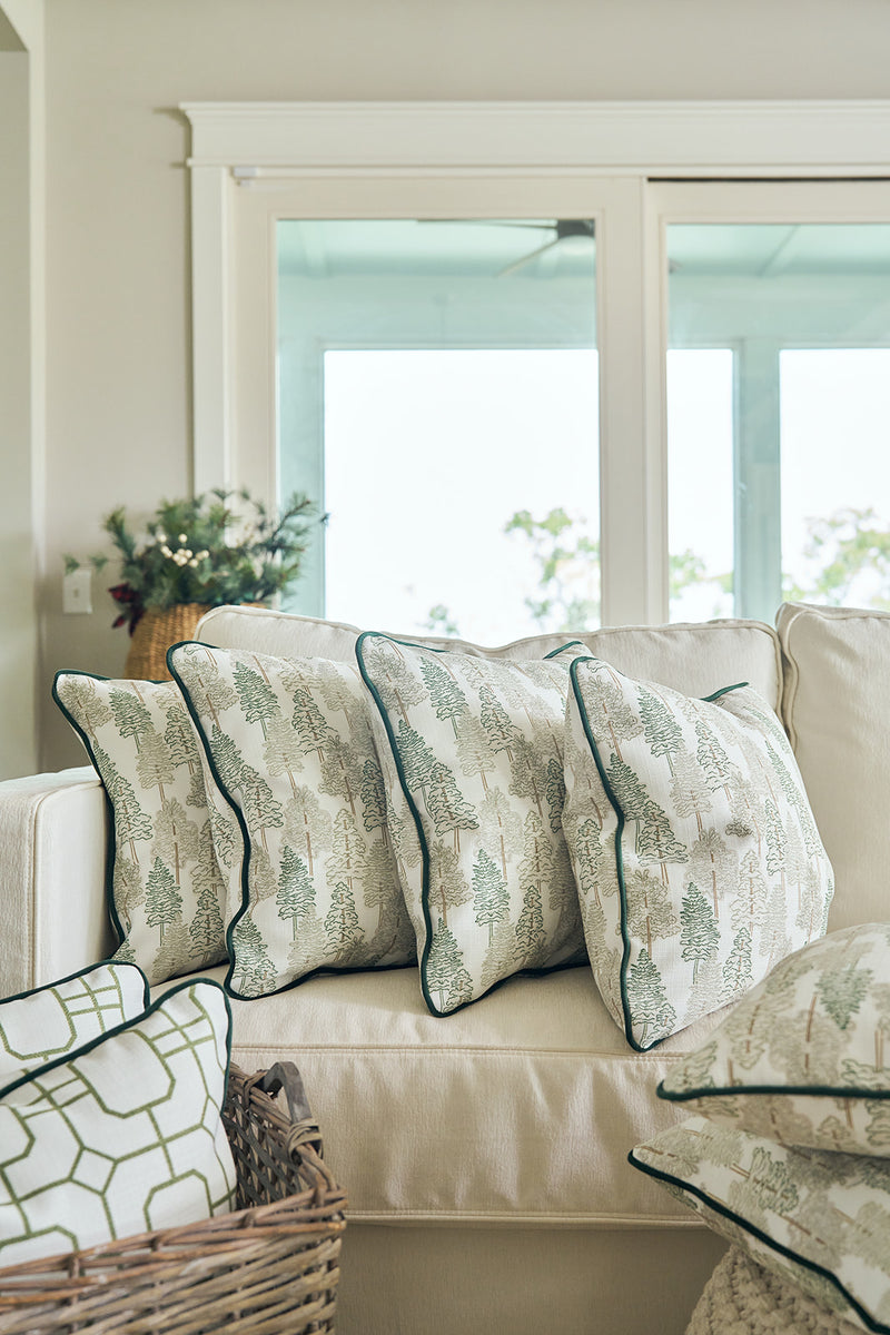 A cozy beige sofa with four pillows, including the Christmas Tree Farm Pillow, sits near a window. A woven basket of extra pillows is in front, while a potted plant brightens the corner.