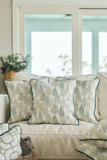 Three Christmas Tree Farm Pillows with dark piping sit on a light-colored sofa in a bright living room, large windows behind and a basket of greenery nearby—perfect for adding festive, holiday charm to your decor.