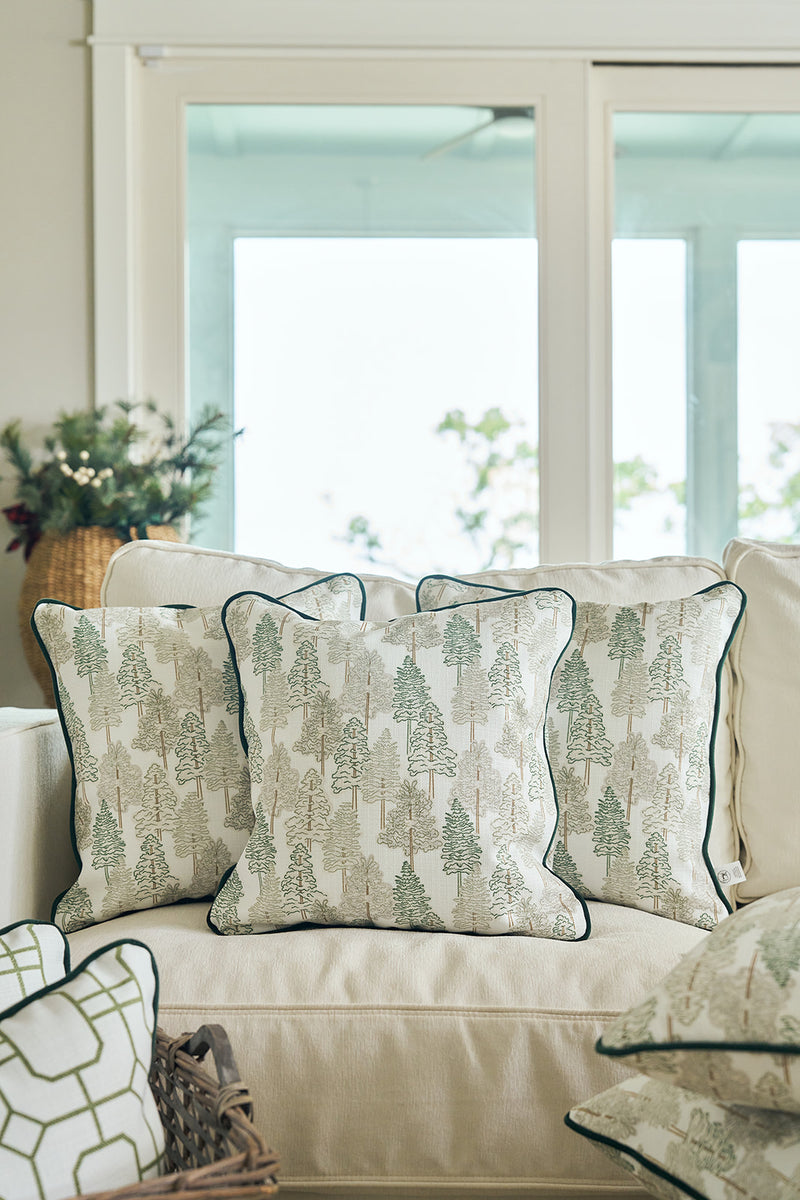 Three Christmas Tree Farm Pillows with dark piping sit on a light-colored sofa in a bright living room, large windows behind and a basket of greenery nearby—perfect for adding festive, holiday charm to your decor.